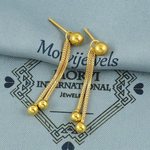 May include: A pair of gold dangle earrings with multiple chains and round beads. The earrings are on a blue fabric background with the text "Morijevels International Jewel"