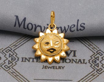 May include: A gold-colored pendant in the shape of a sun with a smiling face. The sun has a detailed design with rays around the face. The pendant is hanging from a small loop. The background is a gray fabric with the word "Morinwels" and "International Jewelry" printed on it.