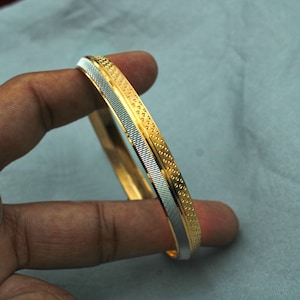 May include: A gold and silver bangle bracelet with a textured design. The bracelet is made of two different metals, with a gold band on the outside and a silver band on the inside.