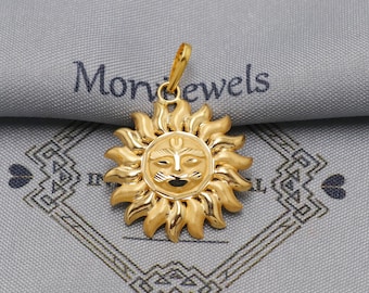 May include: A gold-colored sun pendant with a detailed face and radiating rays. The pendant is suspended from a small loop and is likely made of metal. The background features the text "Monjewels" on a gray fabric.