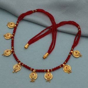 Puede incluir: Un collar de cuentas rojas con colgantes de oro. Los colgantes son circulares con una pequeña piedra roja en el centro. El collar tiene un borla en cada extremo.