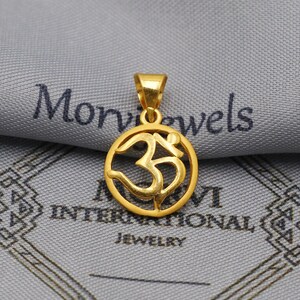 Puede incluir: Un colgante dorado con el símbolo Om dentro de un marco circular. El colgante está suspendido de un pequeño lazo y se encuentra sobre un fondo gris con el texto "Morvi Jewels International Jewelry".