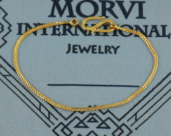 May include: A gold-colored chain bracelet with a hook clasp. The bracelet is displayed on a blue background with the text "MORVI INTERNATIONAL JEWELRY". The chain has a textured, woven design.