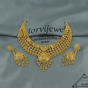 Puede incluir: Un conjunto de joyas de color dorado que incluye un collar y pendientes a juego. El collar presenta un diseño detallado y ornamentado con un colgante central. Los pendientes son de estilo colgante. El nombre de la marca "Morvijejewel" es visible.