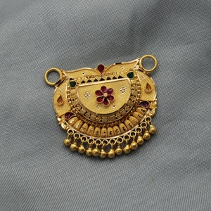 Puede incluir: Colgante dorado ornamentado con un diseño floral, con detalles en rojo y verde. El colgante tiene una superficie detallada y texturizada y pequeñas cuentas doradas colgantes. El fondo es gris liso.
