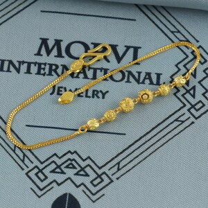 May include: A delicate gold bracelet featuring a series of small, textured floral-shaped beads. The bracelet has a fine chain and a secure clasp. The jewelry is displayed on a blue surface with the text "MOVI INTERNATIONAL JEWELRY".