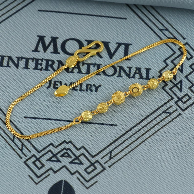 May include: A delicate gold bracelet featuring a series of small, textured floral-shaped beads. The bracelet has a fine chain and a secure clasp. The jewelry is displayed on a blue surface with the text "MOVI INTERNATIONAL JEWELRY".