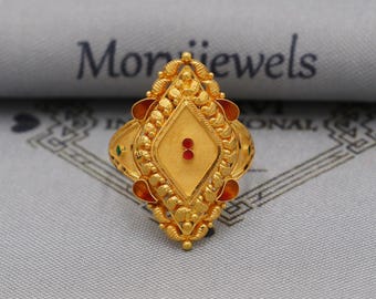 May include: A gold ring with an ornate design. The ring features a central diamond shape with two red dots and is surrounded by intricate detailing. The ring has a textured surface and is set against a gray background.