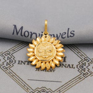 Puede incluir: Un colgante dorado con un diseño de sol con una cara en el centro, rodeado de rayos estilizados. El colgante está suspendido de un pequeño lazo. El fondo incluye el texto "Mon Jewels INTERNATIONAL JEWELRY".