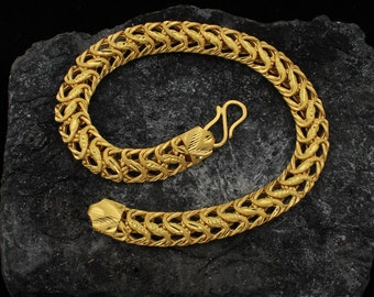 May include: A gold-colored bracelet with a detailed, woven design. The bracelet is made of interlocking links and features a hook clasp. The jewelry is displayed on a dark, textured surface.