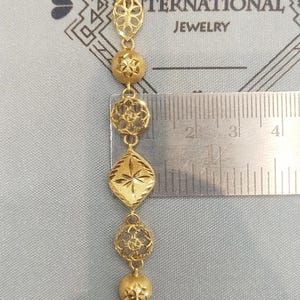 May include: A gold bracelet with intricate designs. The bracelet is made up of a series of connected, ornate, gold pieces. The bracelet is measured against a ruler, showing it is approximately 3 inches long.