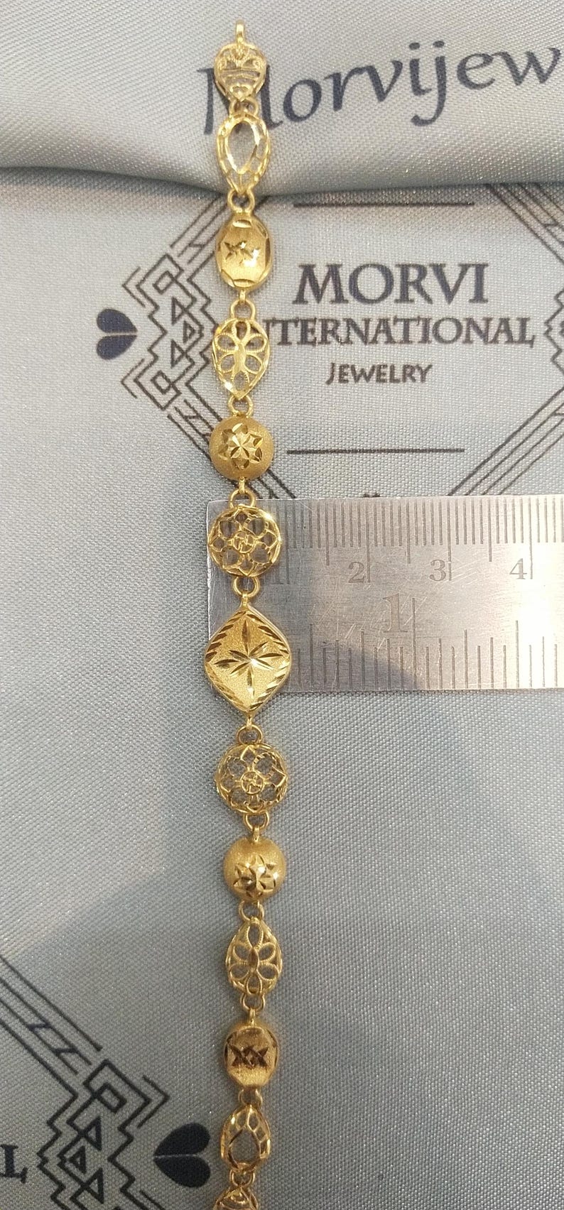 May include: A gold bracelet with intricate designs. The bracelet is made up of a series of connected, ornate, gold pieces. The bracelet is measured against a ruler, showing it is approximately 3 inches long.