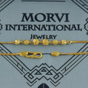 May include: Gold-colored necklace with a decorative chain and a series of round, textured beads. The necklace features a hook clasp and a small, teardrop-shaped end piece. The text "MORVI INTERNATIONAL JEWELRY" is visible in the background.