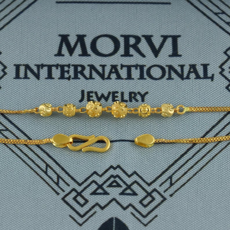May include: Gold-colored necklace with a decorative chain and a series of round, textured beads. The necklace features a hook clasp and a small, teardrop-shaped end piece. The text "MORVI INTERNATIONAL JEWELRY" is visible in the background.