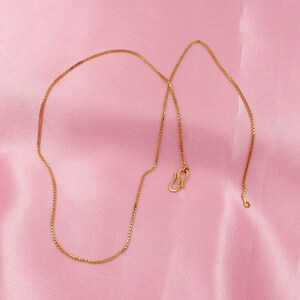 May include: A gold chain necklace with a box chain design. The chain is on a pink background.