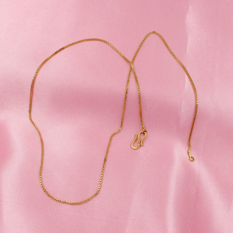 May include: A gold chain necklace with a box chain design. The chain is on a pink background.
