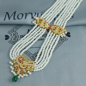 May include: A pearl necklace with multiple strands, featuring gold-coloured clasps and pendants. The clasps and pendants are decorated with red, green, and clear gemstones. A green, teardrop-shaped gemstone dangles from the bottom pendant. The necklace is on a grey surface.