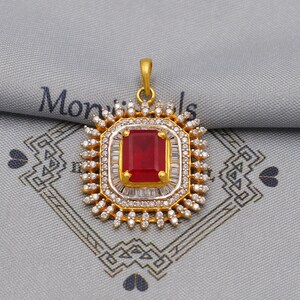 May include: A gold-colored pendant with a large, rectangular, red gemstone at its center. The gemstone is surrounded by smaller, clear stones and a gold frame. The pendant has a loop at the top for a chain. The background is a gray fabric.