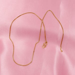 May include: A gold chain necklace with a box chain design. The chain is on a pink background.