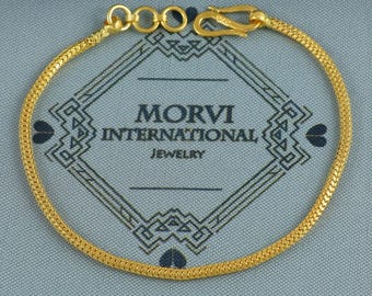 May include: A gold-colored chain bracelet with a hook clasp. The bracelet is circular and lies on a gray surface with the text "MORVI INTERNATIONAL JEWELRY". The bracelet has a textured, woven design.