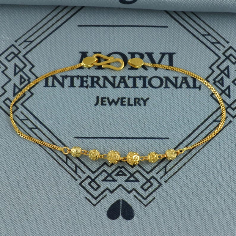 May include: A gold-colored bracelet with a delicate chain and a series of decorative beads. The bracelet features a hook clasp and is displayed against a blue background with the text "HORVI INTERNATIONAL JEWELRY".