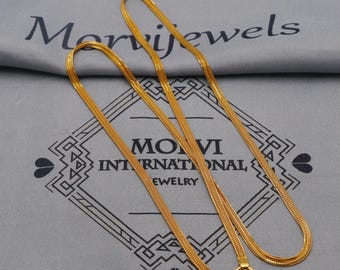 May include: A gold-colored chain necklace with a flat, woven design. The necklace is displayed on a gray surface with the text "Monvi Jewels" and "MONVI INTERNATIONAL JEWELRY". The necklace has a hook clasp.