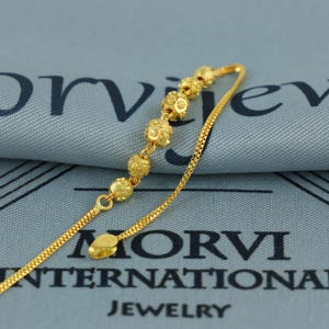 May include: A gold-colored necklace with a delicate chain and a series of decorative, spherical beads. The beads have a textured surface. The necklace is displayed on a blue fabric with the text "MORVI INTERNATIONAL JEWELRY".