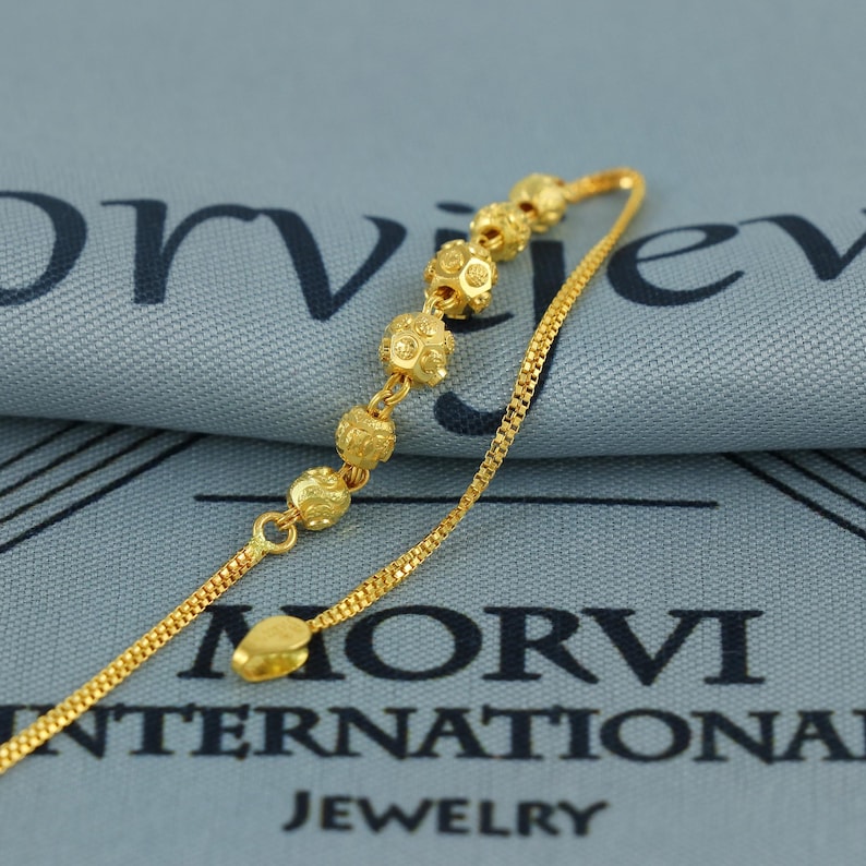 May include: A gold-colored necklace with a delicate chain and a series of decorative, spherical beads. The beads have a textured surface. The necklace is displayed on a blue fabric with the text "MORVI INTERNATIONAL JEWELRY".