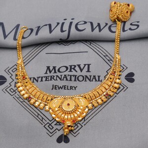 Op de afbeelding: Een goudkleurige ketting- en ringenset. De ketting heeft een gebogen ontwerp met ingewikkelde details en kleine rode accenten. De ring heeft een bijpassend ontwerp. De tekst "MORVI INTERNATIONAL JEWELRY" is zichtbaar.