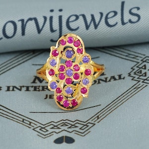 Könnte beinhalten: Ein goldfarbener Ring mit einem kunstvollen Design, besetzt mit lila und rosa Edelsteinen. Der Ring hat ein detailliertes filigranes Muster, und die Edelsteine sind rund geschliffen. Der Ring ist vor einem blauen Hintergrund platziert.