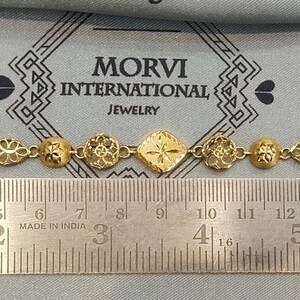 May include: A gold bracelet with intricate designs, measuring approximately 7 inches long. The bracelet is made of small, connected pieces with a central diamond-shaped piece. The bracelet is displayed on a ruler with the text "MORVI INTERNATIONAL JEWELRY" visible above it.