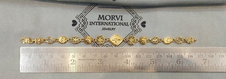 May include: A gold bracelet with intricate designs, measuring approximately 7 inches long. The bracelet is made of small, connected pieces with a central diamond-shaped piece. The bracelet is displayed on a ruler with the text "MORVI INTERNATIONAL JEWELRY" visible above it.