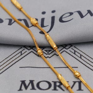 Può includere: Primo piano di due collane d'oro con maglie intricate e perline decorative. Le collane sono esposte su una superficie grigia con la parola "MORVI" visibile. Le catene hanno un aspetto strutturato e sono probabilmente in oro.