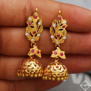 Puede incluir: Un par de pendientes dorados con intrincados diseños florales y de hojas. Los pendientes presentan piedras preciosas rojas y blancas, con una base en forma de campana. Los pendientes cuelgan de una pieza superior decorativa.