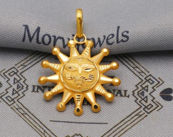 May include: A gold-colored sun pendant with a smiling face and rays. The pendant is suspended from a small loop. The background is a gray fabric with the text "Mon jewels" visible.