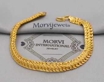 May include: A gold-colored bracelet with a textured, woven design. The bracelet is displayed on a gray surface with the text "Morvijewels" and "MORVI INTERNATIONAL JEWELRY". The bracelet has a hook clasp.