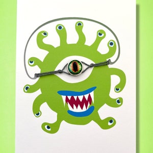 Könnte beinhalten: Eine weiße Karte mit einem grünen Monster mit mehreren Augen und einem breiten Grinsen. Das Monster trägt ein graues Armband aus Schnur mit einem grünen und schwarzen Augen-Anhänger. Auf der Karte steht "Armbänder nicht im Lieferumfang enthalten! Bitte in gewünschter Farbe separat bestellen."