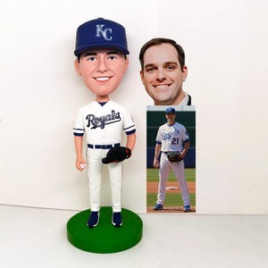 Könnte beinhalten: Eine Bobblehead-Figur eines Baseballspielers in einem weiß-blauen Royals-Trikot und einer Kappe. Die Figur hält einen Baseball und einen schwarzen Handschuh. Der Wackelkopf steht auf einer grünen Basis.