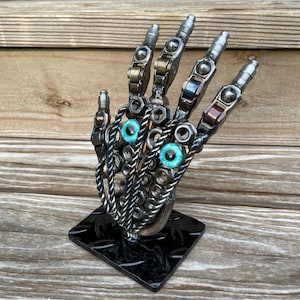 May include: A metal sculpture of a hand with turquoise-colored eyes. The fingers and palm are constructed from various metal pieces, including nuts, bolts, and other hardware. The hand is mounted on a black, diamond-plate base.