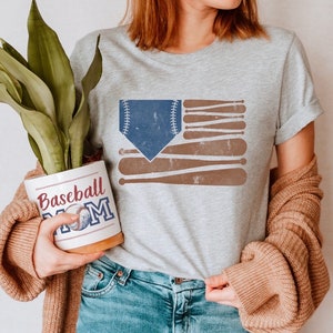 May include: A woman wearing a grey t-shirt with a baseball-themed American flag design. The shirt says "Baseball Mom". She is holding a white ceramic planter with the same design.