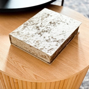 以下が含まれることがあります： 明るい茶色の丸い木製コーヒーテーブル。上には質感のある明るい色の箱が置かれています。テーブルの側面にはリブのデザインがあります。背景には黒いテーブルが見えます。