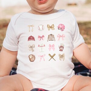 May include: A white baby bodysuit adorned with baseball-themed illustrations in pink and brown, including bows, a baseball, a helmet, a cap, a glove, and crossed bats. The baby is sitting on a plaid blanket.