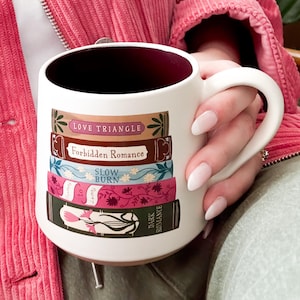 May include: White ceramic mug with a stack of books design. Each book has a different title: "Love Triangle", "Forbidden Romance", "Slow Burn", and "Dark Romance". The mug has a dark burgundy interior and a large handle.