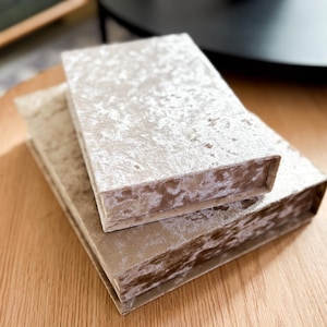 以下が含まれることがあります： 木製のテーブルに積み重ねられた2つの長方形の箱。箱は、質感のある明るいベージュのベルベット素材で覆われています。箱はサイズが異なり、小さい方が上にあります。