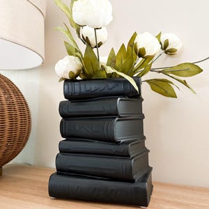 May include: A black ceramic vase in the shape of stacked books, filled with white flowers and green foliage. The vase is on a wooden surface, with a wicker lamp in the background.
