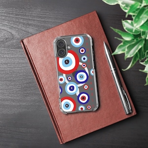 Puede incluir: Una funda transparente para teléfono con un fondo gris que presenta un patrón de diseños de ojos rojos, blancos y azules. La funda está sobre un cuaderno cerrado de cuero marrón. Un bolígrafo plateado descansa junto al cuaderno.