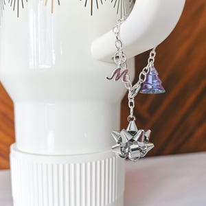 May include: A silver charm bracelet with three dangling charms: a letter "M", a purple Christmas tree, and a silver bow. The bracelet is hanging on a white mug with a gold design.