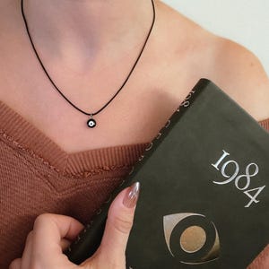 Peut inclure: Un collier en cordon noir avec un petit pendentif rond représentant un œil bleu et blanc. Le livre "1984" est tenu dans la main, avec le titre et un symbole d'œil visibles sur la couverture. La personne porte un pull marron.