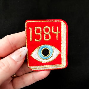 May include: A red embroidered patch in the shape of a book with the number "1984" in gold lettering. The patch features a blue eye with a black pupil and white accents. The patch has a gold border.