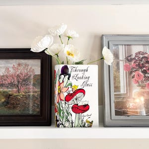May include: A decorative shelf arrangement featuring a vase shaped like a book with the text "Through the Looking Glass" and a colorful mushroom illustration. The vase holds white flowers. Flanking the vase are framed pictures.